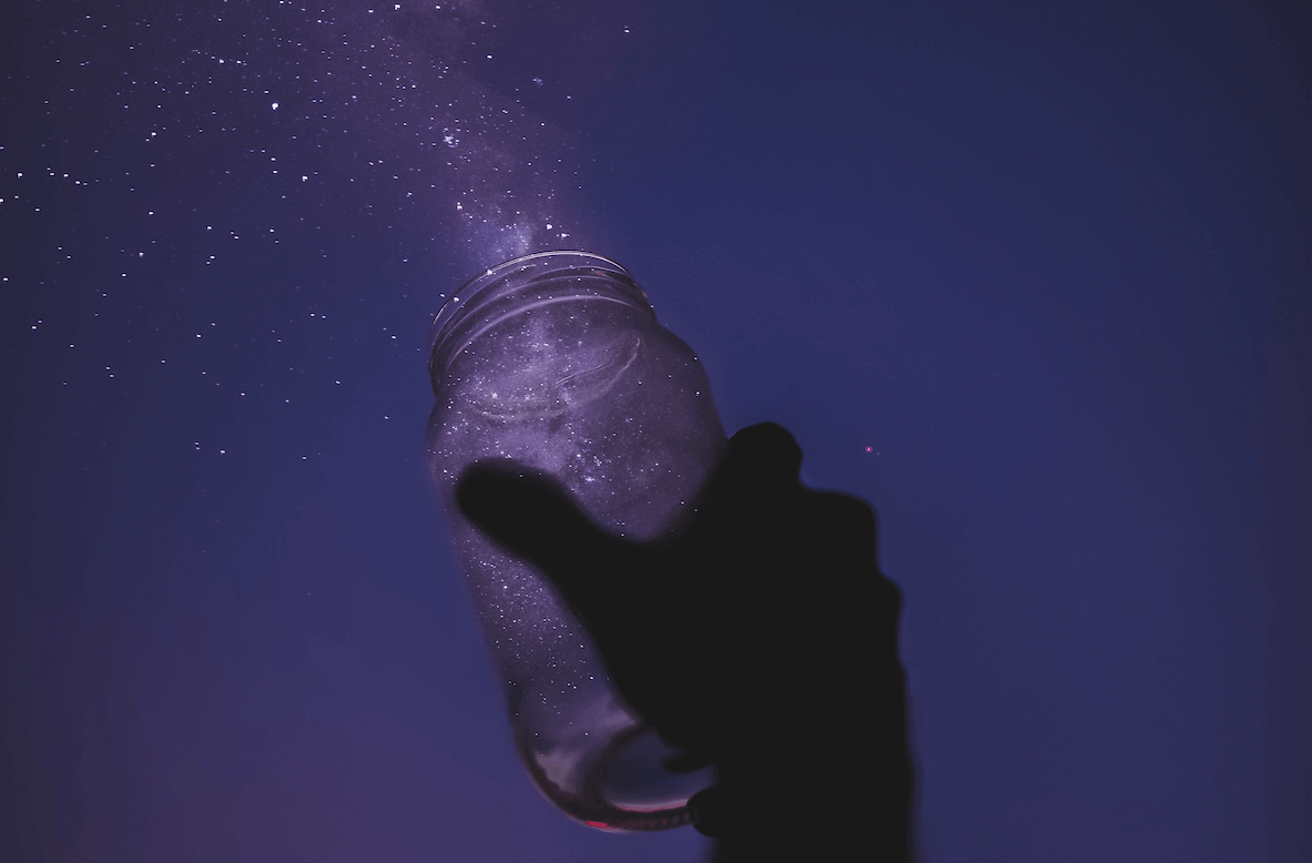 A hand holding a glass jar and letting out golden dust to look like "magic" with a purple sky